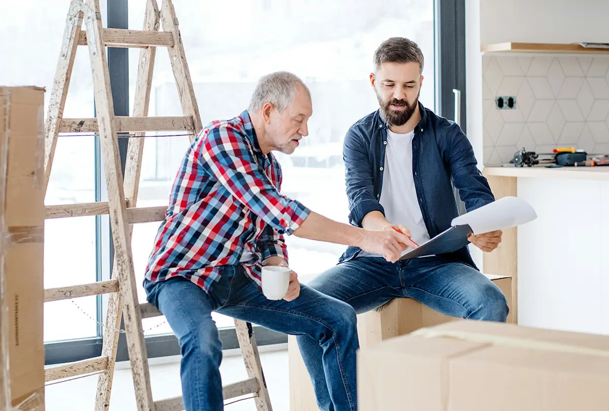 Two people are sitting in a new home, discussing and looking over compliance papers on a clipboard together.